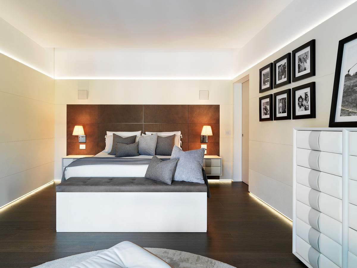 Camera da letto con illuminazione perimetrale, letto imbottito con panca imbottita e comodini sospesi, in atmosfera raccolta