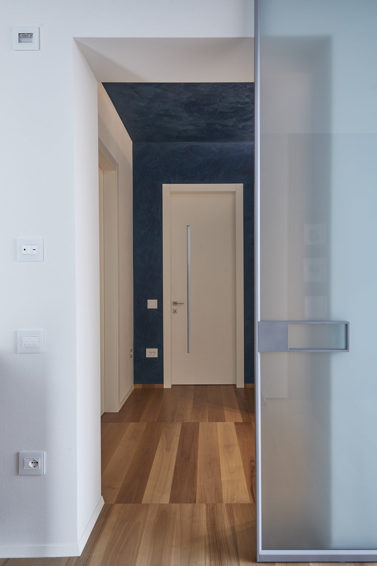 Ingresso interno con porta moderna bianca, nicchia blu e pavimento in legno