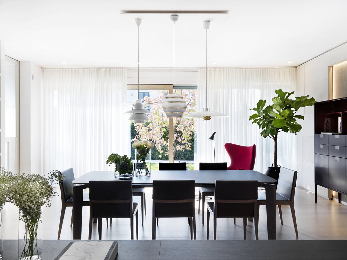 Vista della zona pranzo con tavolo in legno a 8 posti, lampade sospese di design e finestre scorrevoli con tende a tutta altezza e scorcio del giardino esterno
