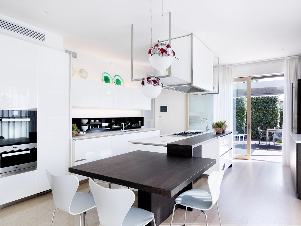Cucina bianca con isola e zona pranzo integrata, elementi sospesi, sospensioni in vetro di design e vista sullo spazio pranzo outdoor