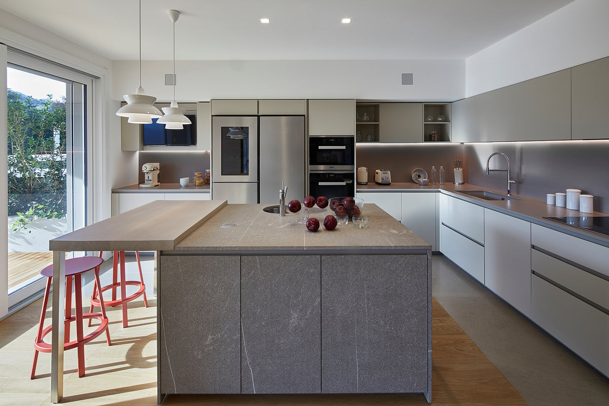 Cucina con grande isola conviviale in pietra fiammata, colonne integrate, elettrodomestici professionali in acciaio inox e finiture neutre