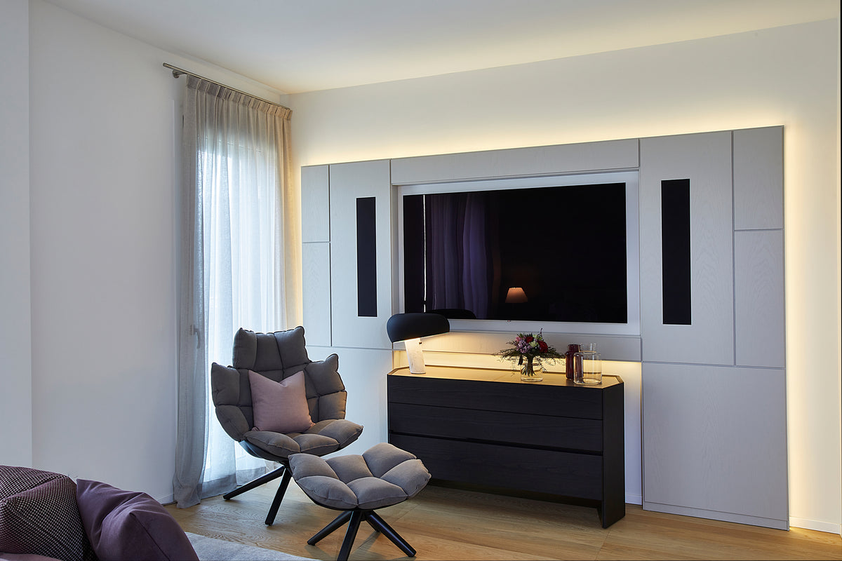 Angolo relax in zona notte con poltrona imbottita e pouf, madia in legno scuro e parete TV integrata