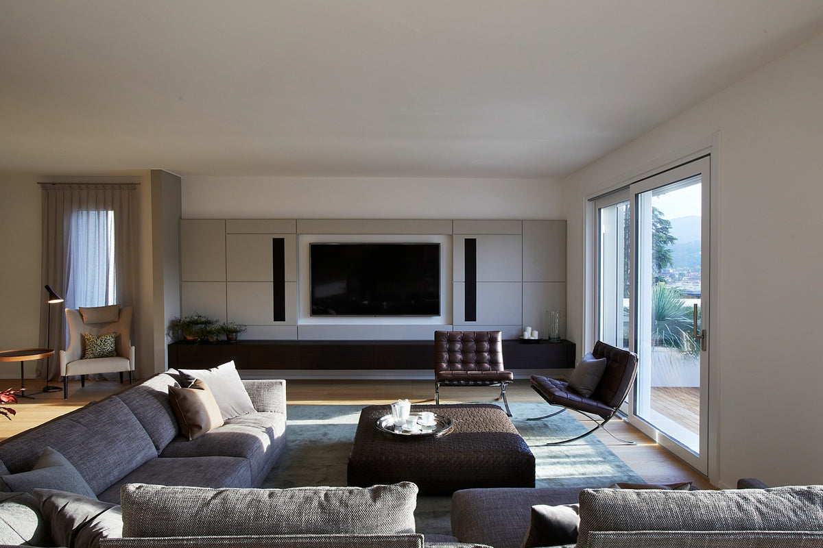 Living con grande divano modulare, parete TV decorativa, poltrone di design in pelle e vetrata verso l’esterno, in progetto residenziale su misura