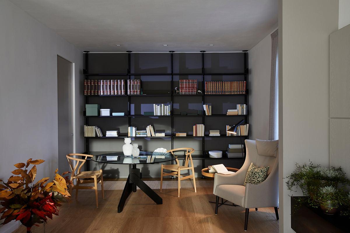 Angolo raccolto del living con zona lettura con libreria a parete, tavolo rotondo in vetro e sedute in legno e poltrona in pelle