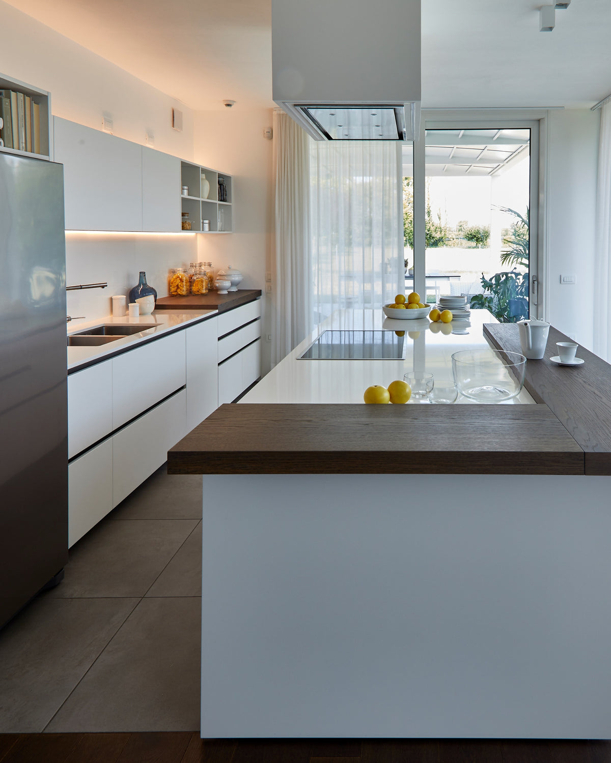 Dettaglio cucina bianca moderna con isola snack in legno scuro, vetrata con vista sulla zona living ourdoor