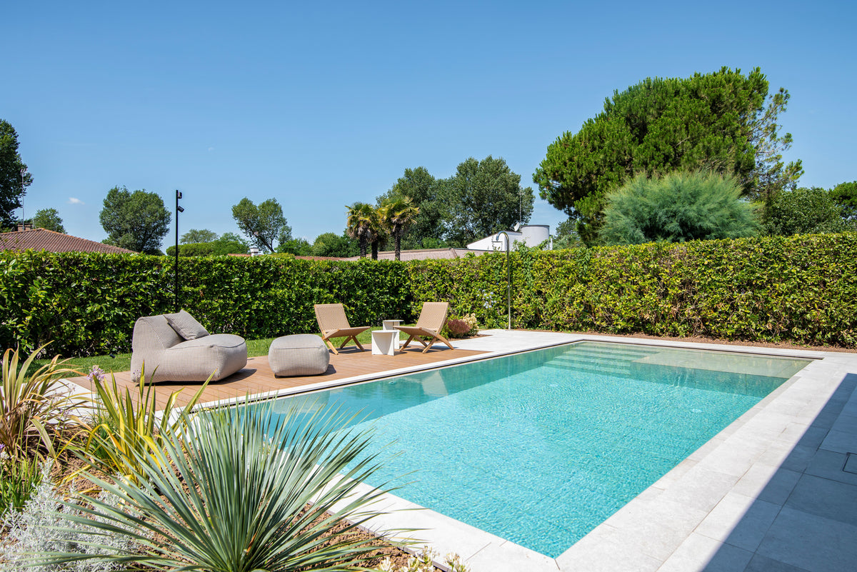 Piscina e area relax outdoor con pavimentazione chiara, deck in legno e giardino piantumato