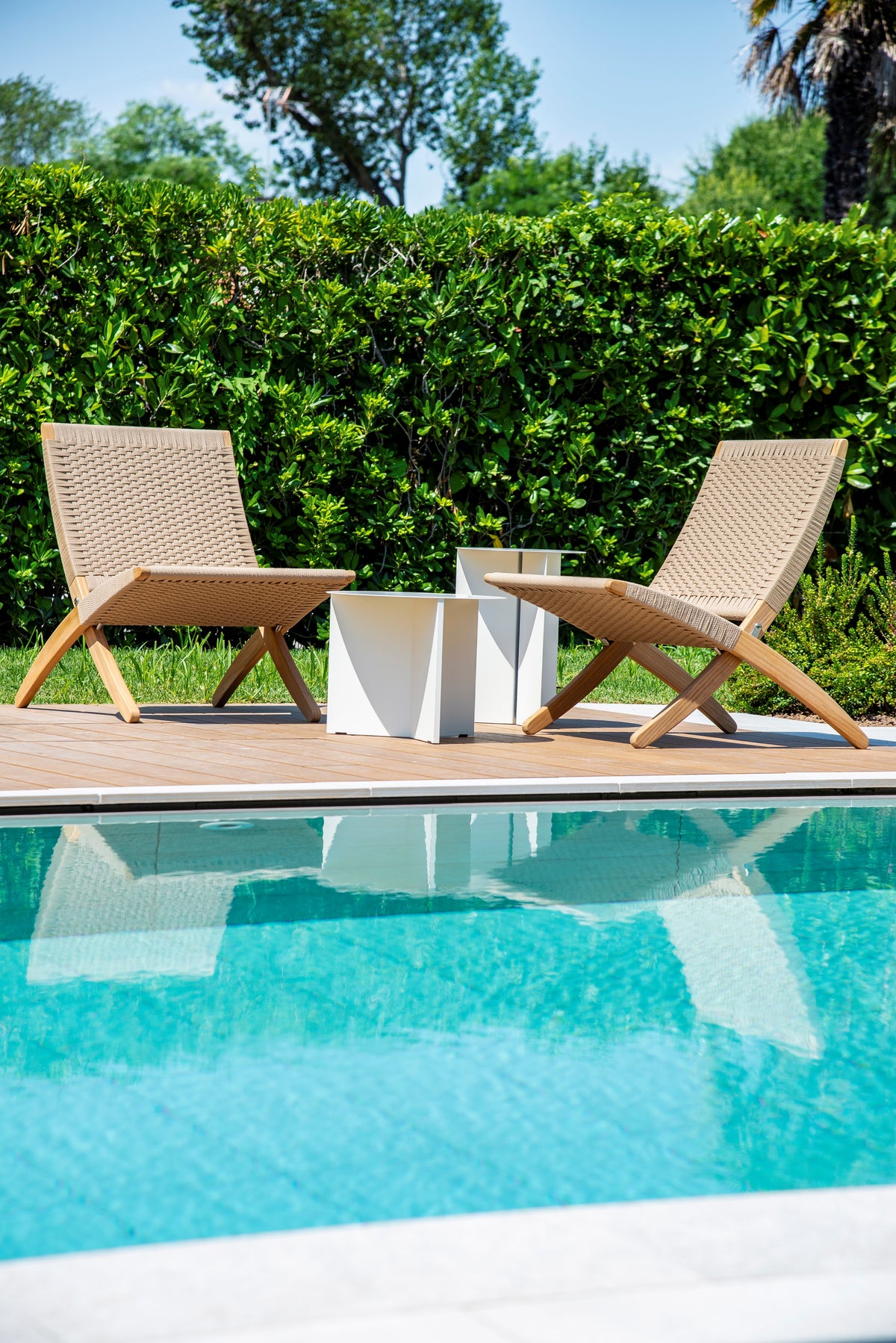 Sedute outdoor in legno a bordo piscina con tavolini e siepe verde sullo sfondo