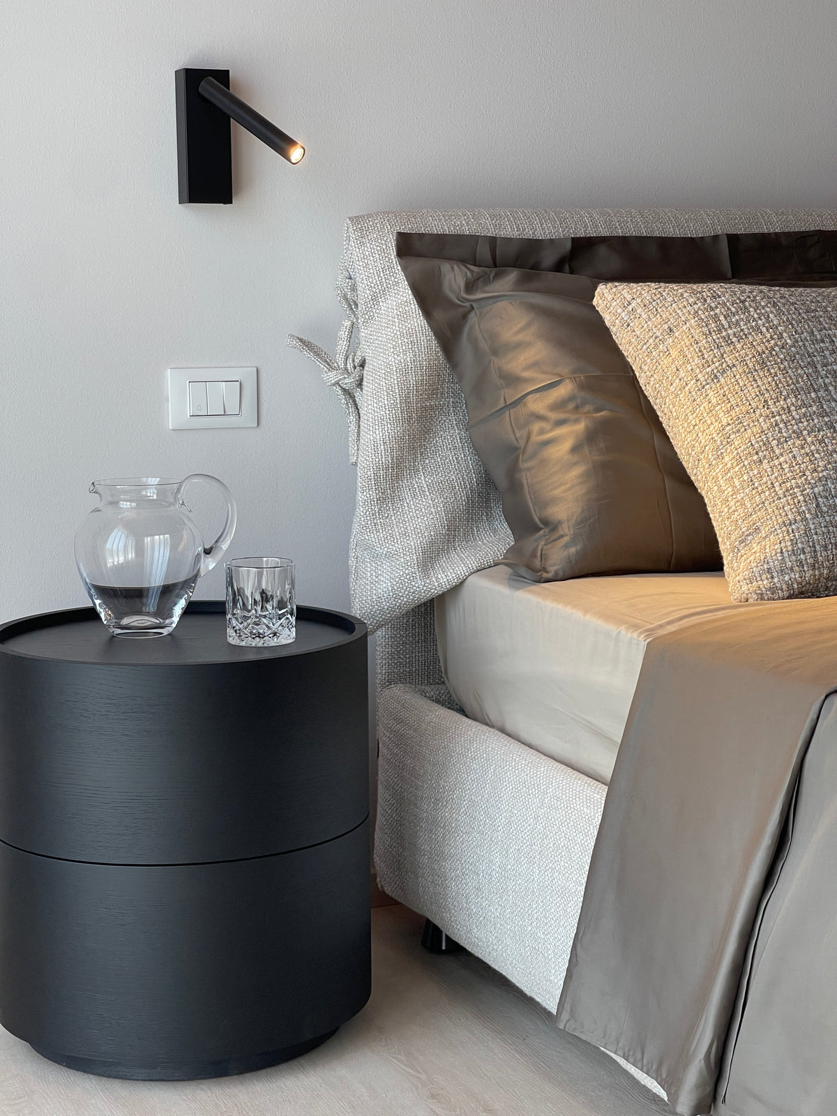 Dettaglio camera da letto moderna con comodino nero, applique orientabile e testiera tessile