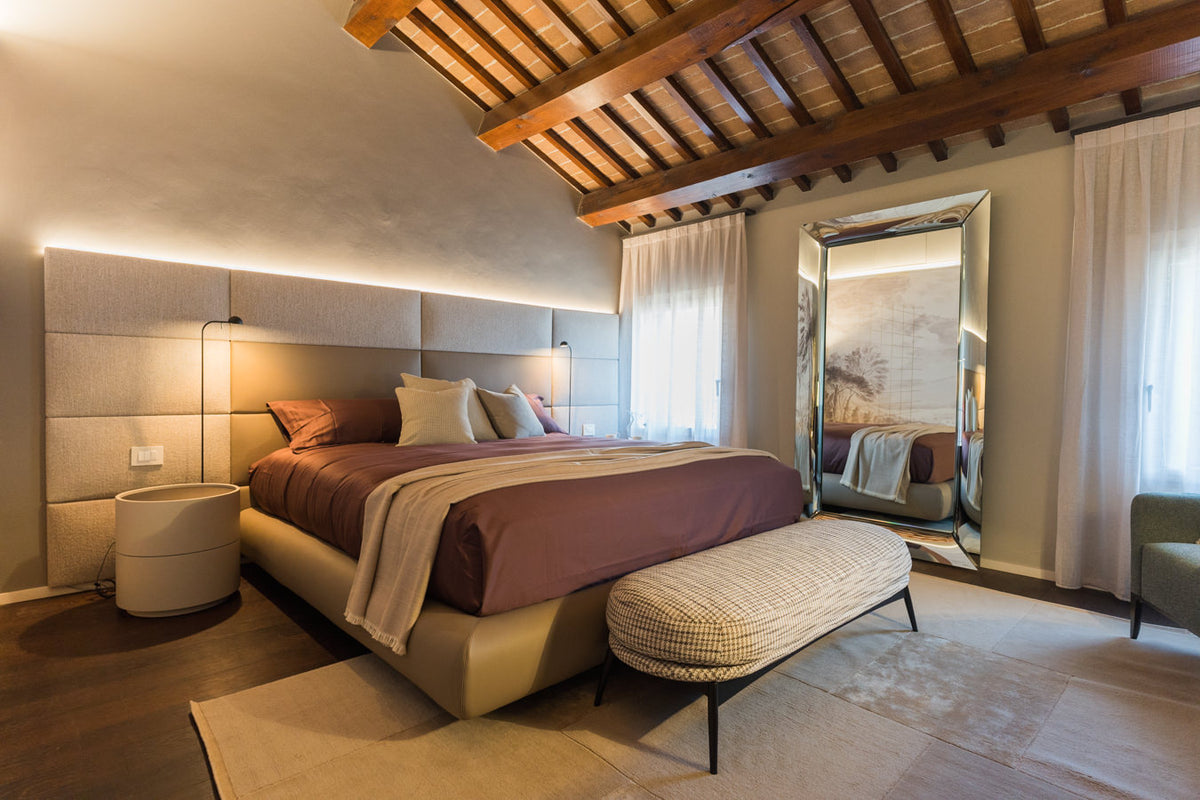 Camera padronale con letto imbottito, boiserie tessile illuminata e travi in legno a vista, dalle finiture in equilibrio tra classico e contemporaneo