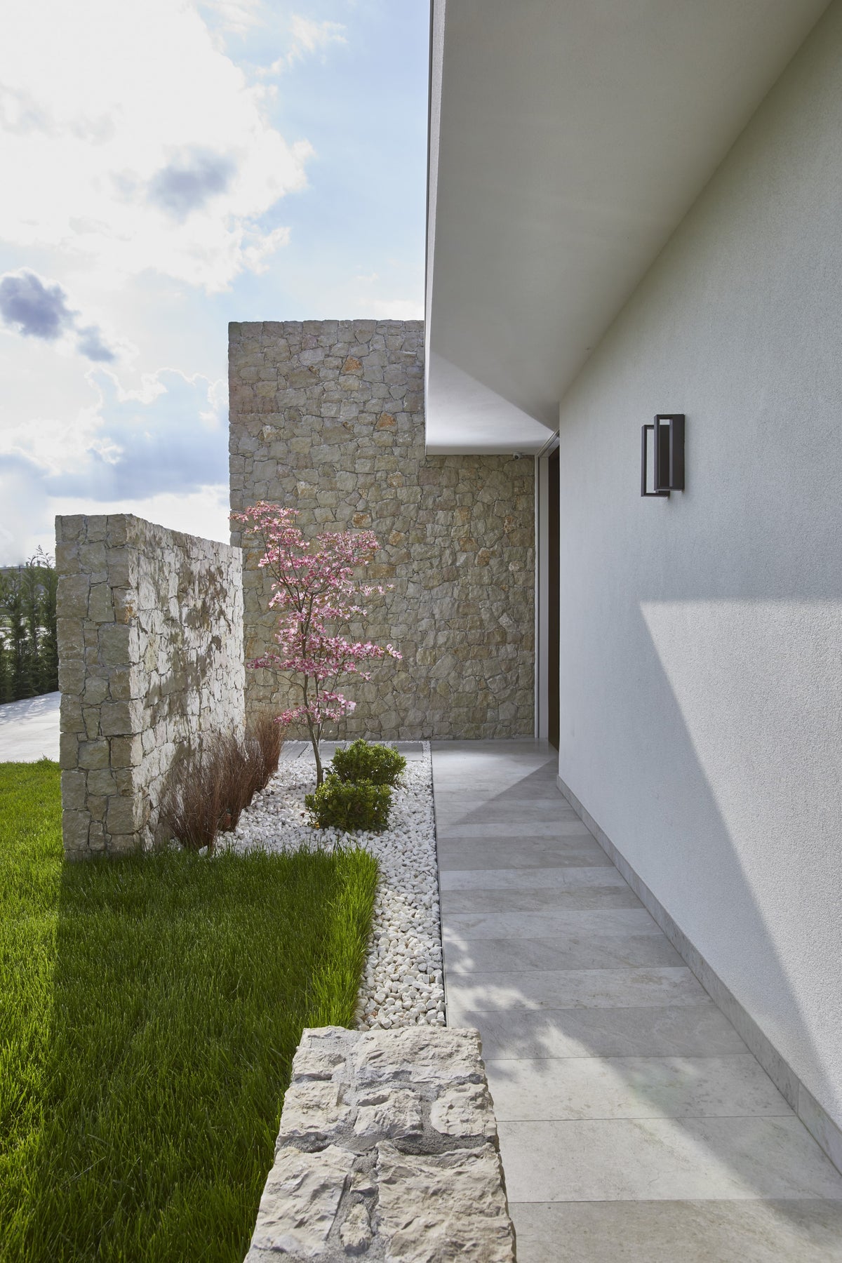 Percorso esterno della villa con parete in pietra, prato e volumi architettonici essenziali