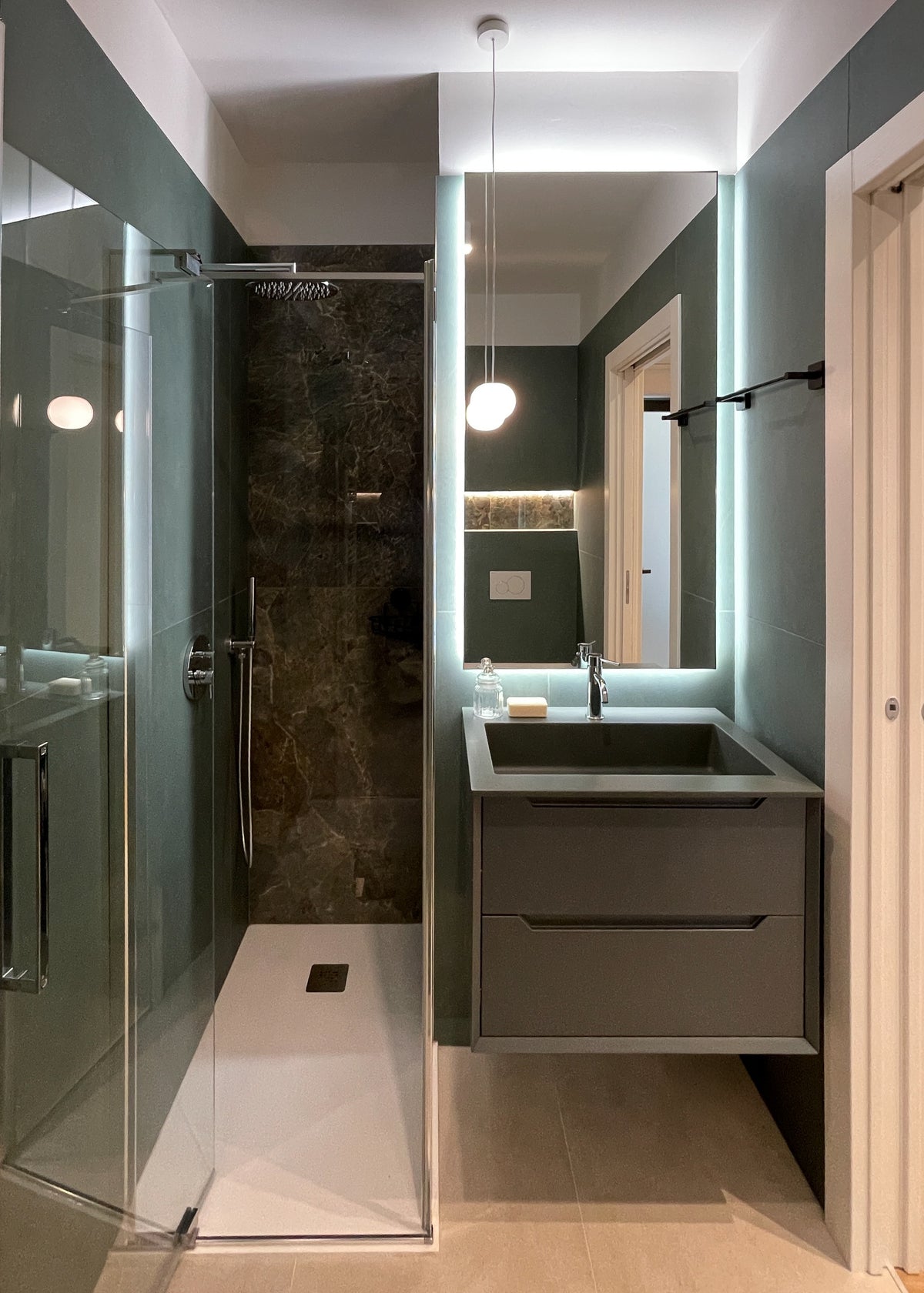 Progettazione bagno contemporaneo con doccia, mobile sospeso, illuminazione integrata e superfici materiche