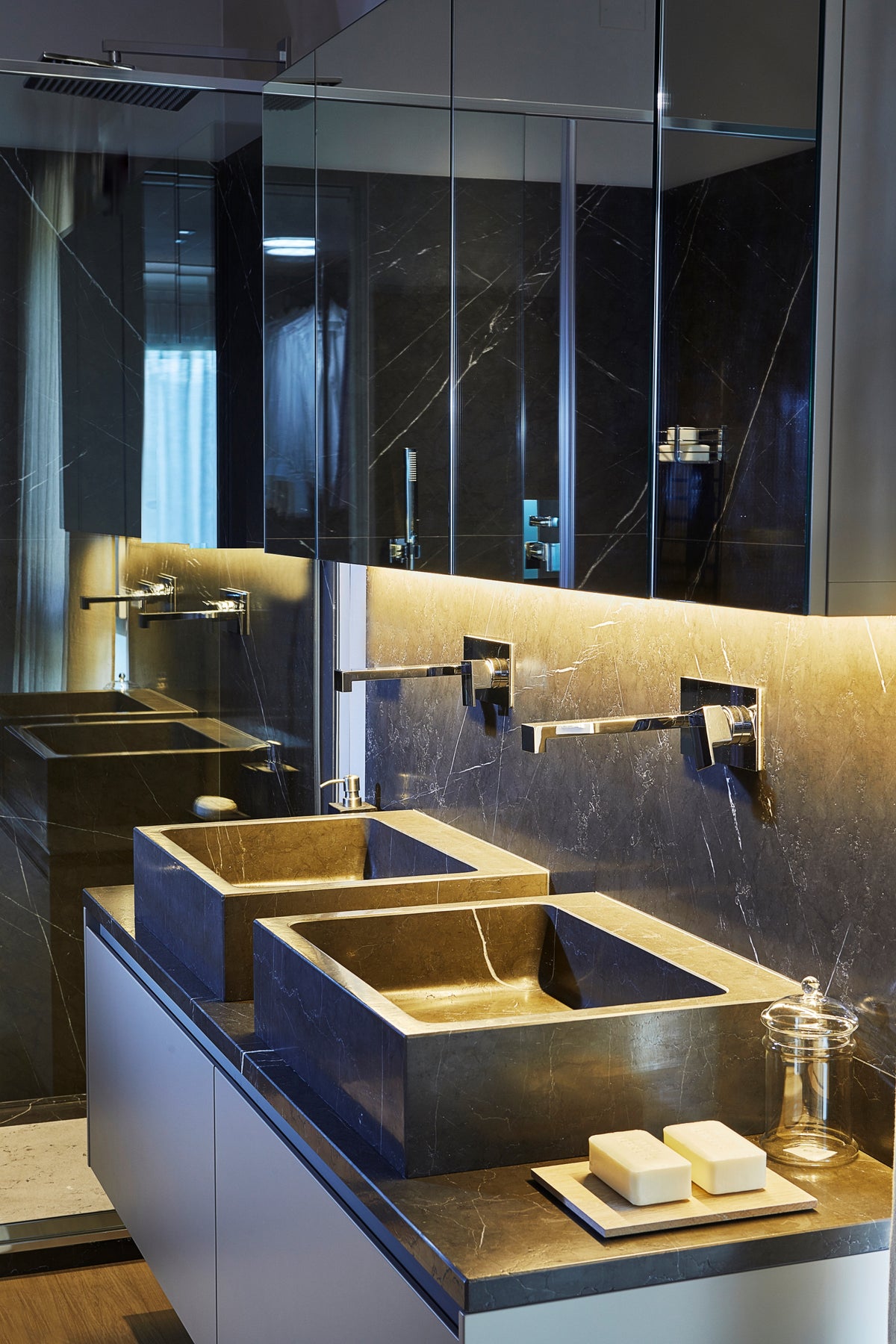 Bagno con doppio lavabo in pietra scura, pensili contenitori con specchi e rivestimenti materici