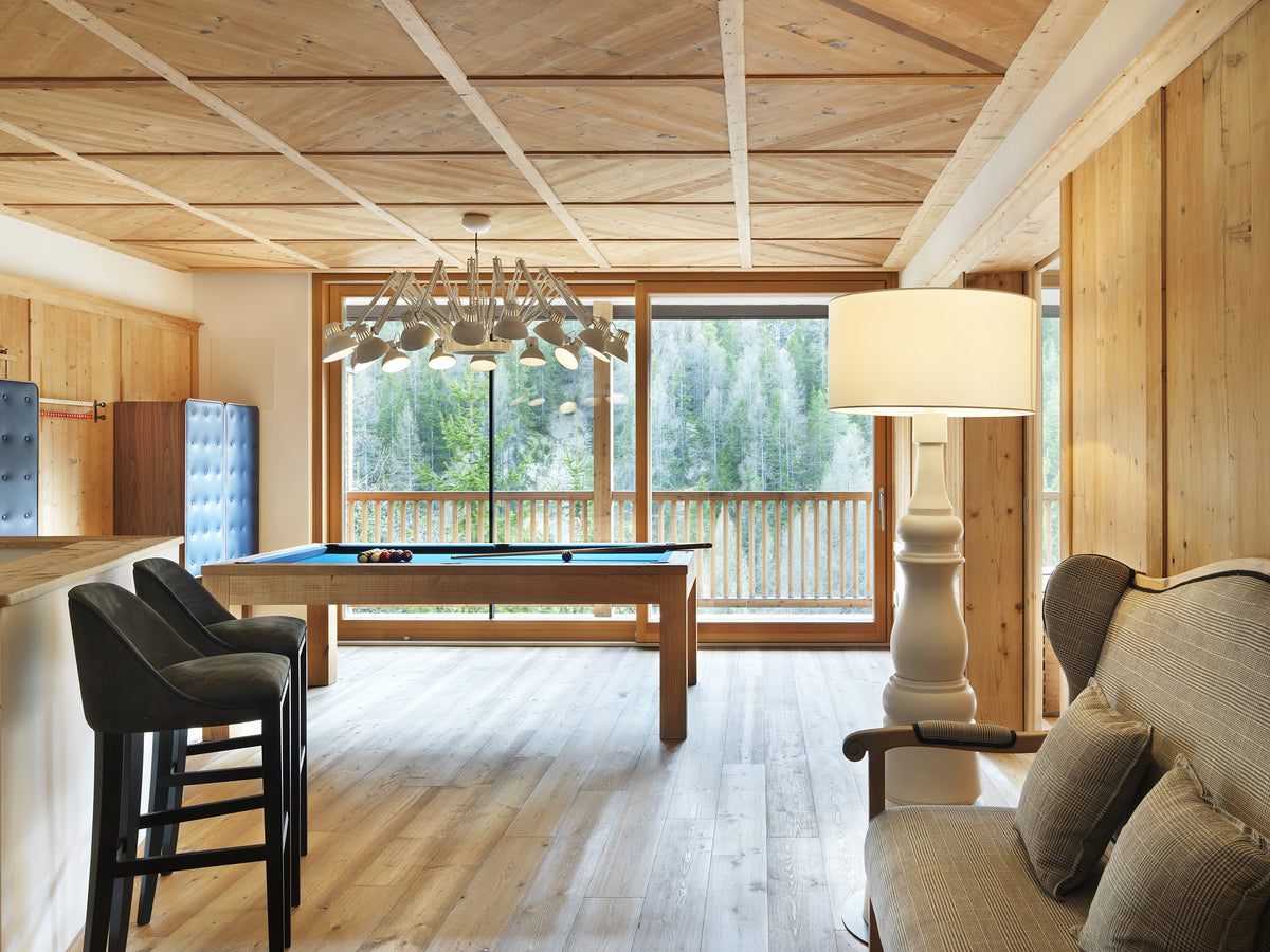 Sala biliardo con bancone bar, soffitto in legno, lampadario scenografico e grande vetrata sul paesaggio alpino