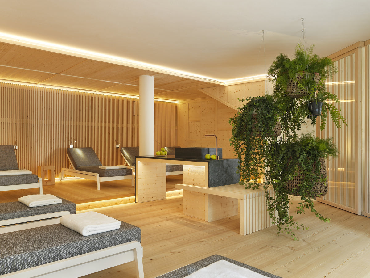 Lounge benessere con lettini imbottiti, bancone centrale e linee morbide in legno chiaro, con giardino sospeso
